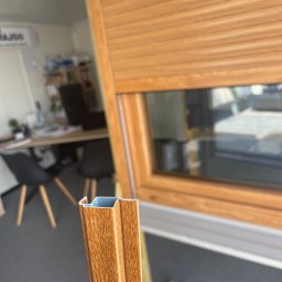 Majgo Łukasz Rudzki - Przekrój profilu okiennego w kolorze drewna na tle okna z zewnętrzną roletą, widoczne biuro w tle.