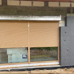 Majgo Łukasz Rudzki - Zamknięta zewnętrzna roleta w kolorze jasnego drewna, zamontowana w oknie na surowym etapie budowy, widoczne ocieplenie budynku i folia ochronna na ramie okiennej.