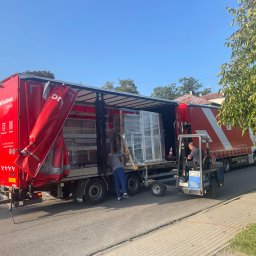Majgo Łukasz Rudzki - Rozładunek okien PCV z ciężarówki za pomocą wózka widłowego na tle ulicy i drzew.