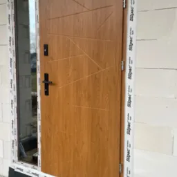 Drewnopodobne drzwi zewnętrzne z przeszkleniem bocznym, zabezpieczone taśmami montażowymi na surowej ścianie z betonu komórkowego.