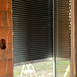 Majgo Łukasz Rudzki - Montaż zewnętrznej rolety okiennej w surowym stanie, widoczna drabina, mur z cegły i wystające kable.