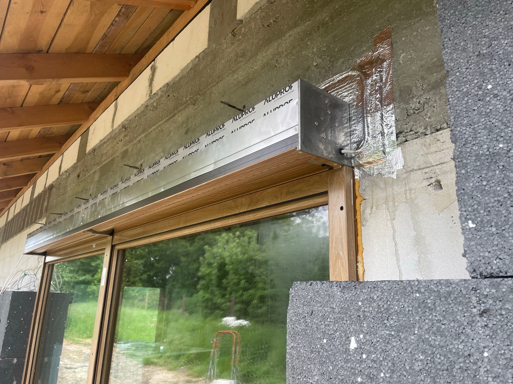 Montaż zewnętrznej rolety z aluminiową obudową marki Aluprof nad oknem z drewnianą ramą, widoczne elementy konstrukcyjne budynku i warstwa izolacyjna