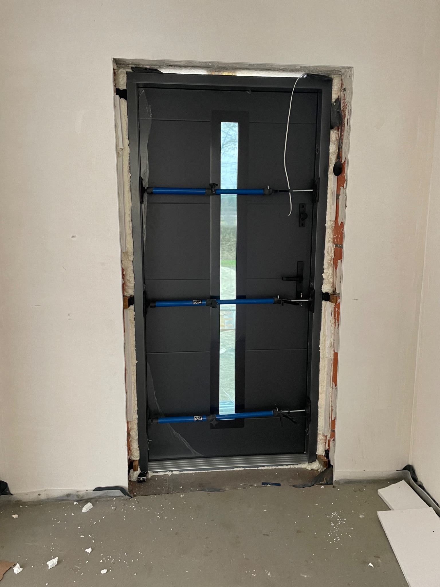 Montaż nowoczesnych, grafitowych drzwi wejściowych z pionowym przeszkleniem, zabezpieczonych rozpórkami montażowymi, widoczne pianka montażowa i fragmenty cegły w ościeżnicy.