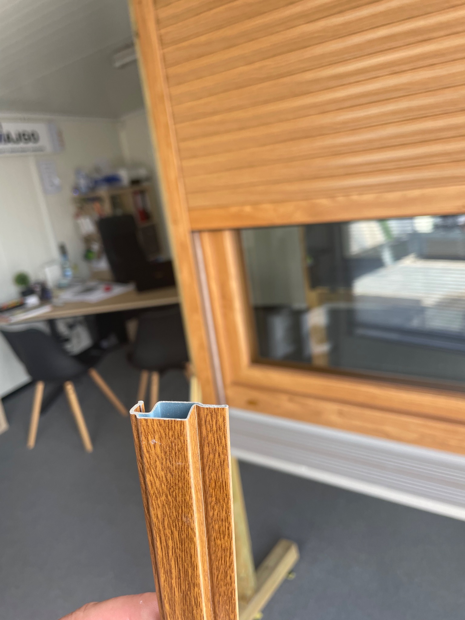 Przekrój profilu okiennego w kolorze drewna na tle okna z zewnętrzną roletą, widoczne biuro w tle.