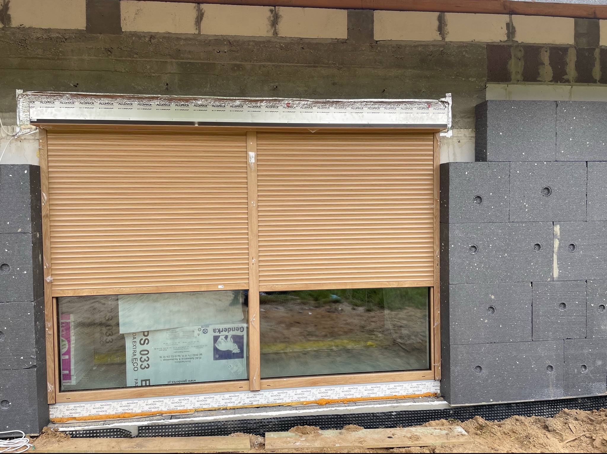 Zamknięta zewnętrzna roleta w kolorze jasnego drewna, zamontowana w oknie na surowym etapie budowy, widoczne ocieplenie budynku i folia ochronna na ramie okiennej.