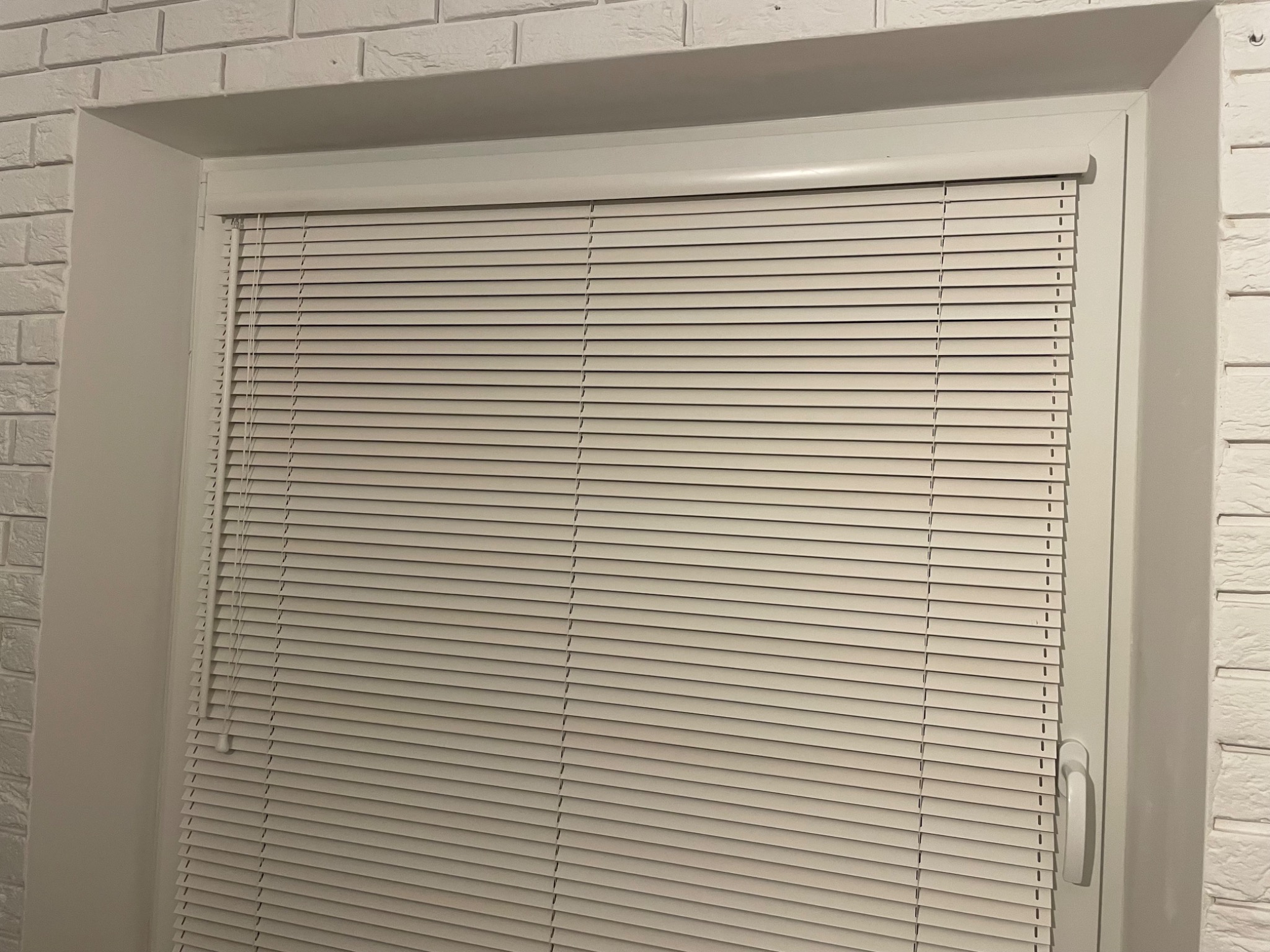 Zamknięte jasne żaluzje poziome w oknie z białymi ramami, widoczne na tle ściany imitującej białą cegłę.