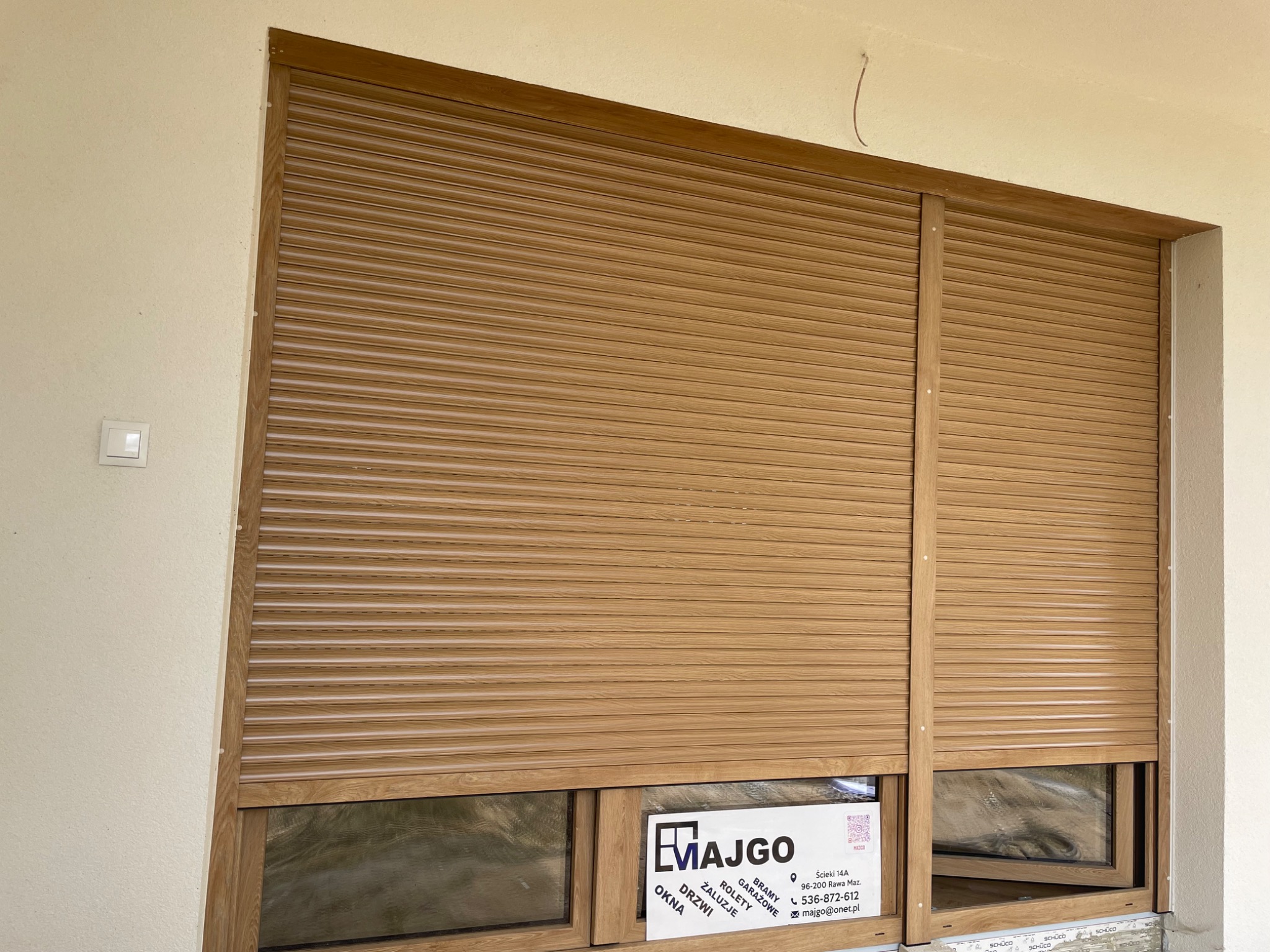 Zewnętrzne rolety w kolorze jasnego drewna, częściowo opuszczone, zamontowane na oknie. Widoczna naklejka firmy na ramie okiennej.