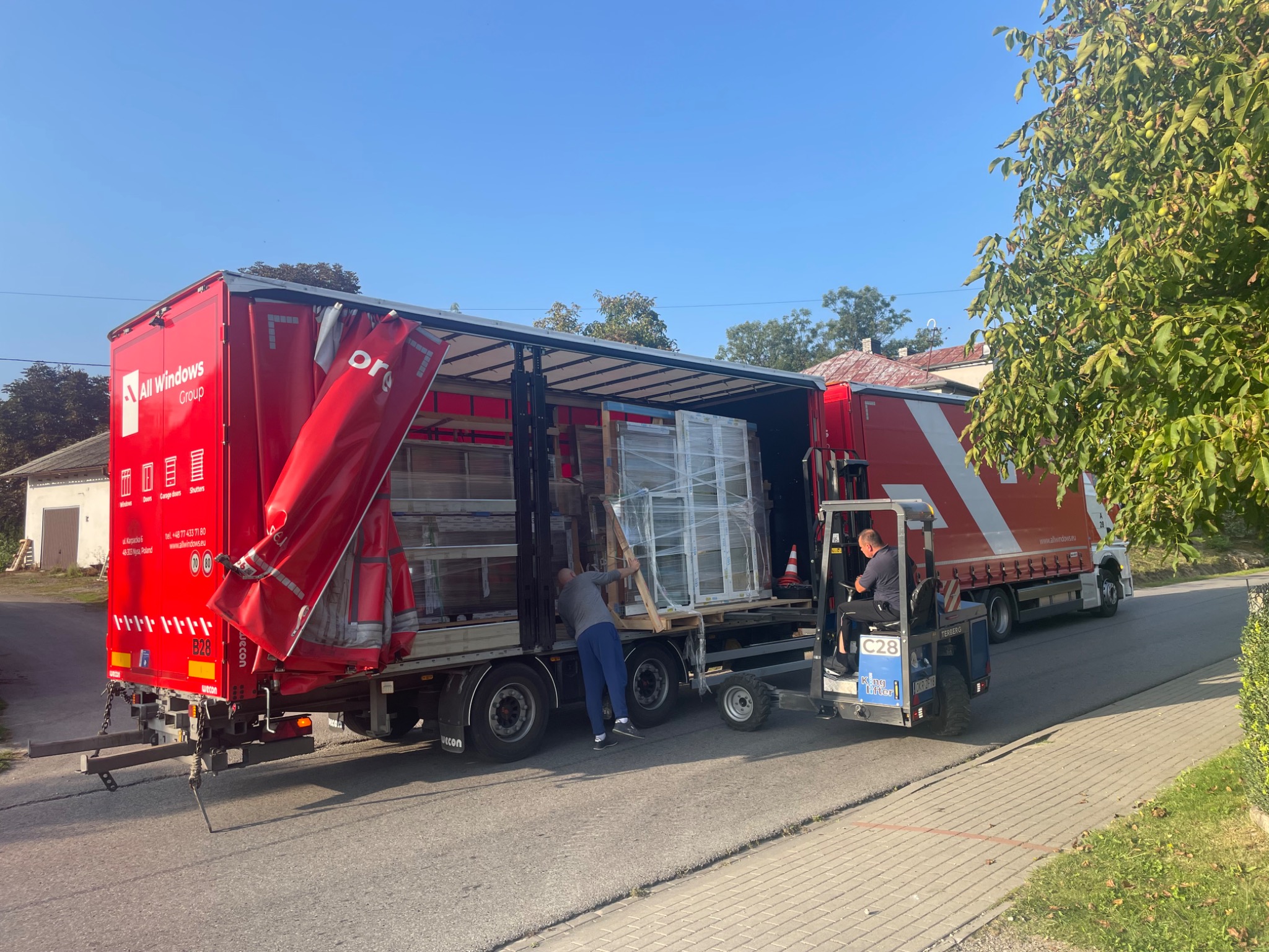Rozładunek okien PCV z ciężarówki za pomocą wózka widłowego na tle ulicy i drzew.