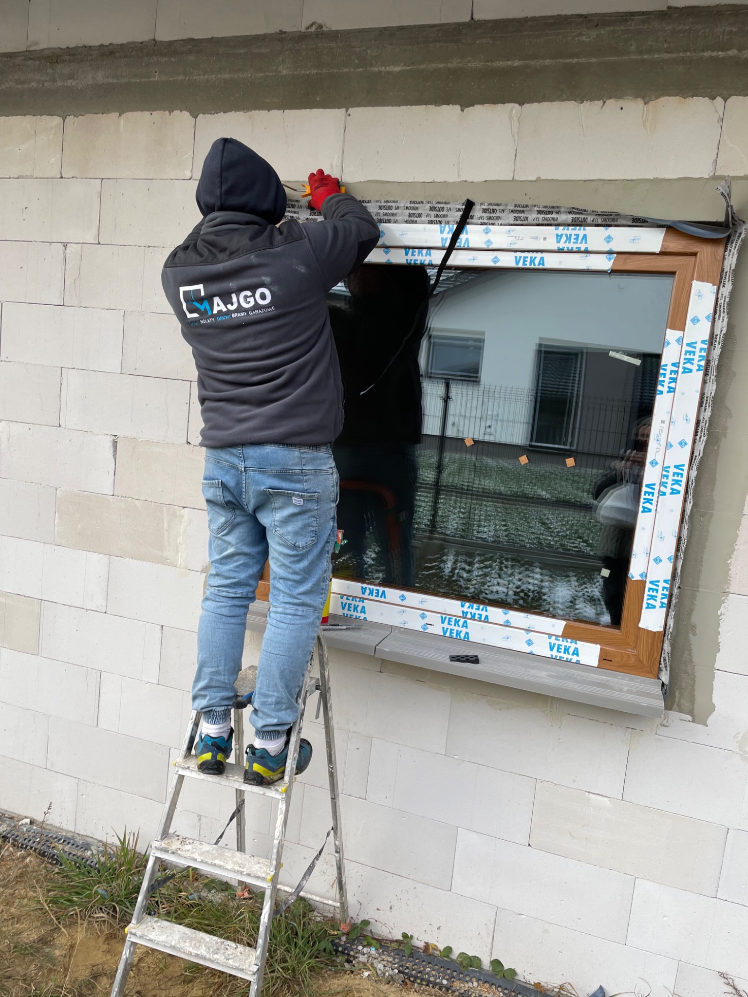 Montera w bluzie z logo firmy na drabinie, uszczelnia brązowe okno w surowym murze z betonu komórkowego, widoczne taśmy uszczelniające marki VEKA.