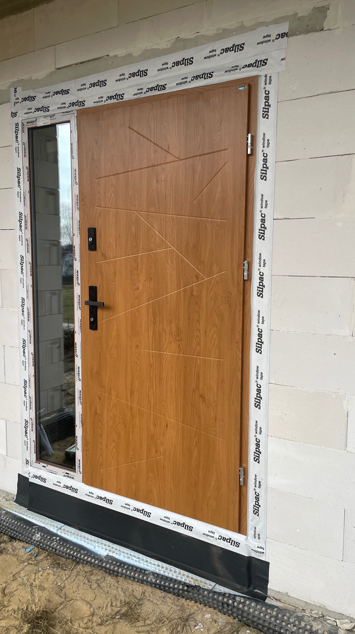 Drewnopodobne drzwi zewnętrzne z przeszkleniem bocznym, zabezpieczone taśmami montażowymi na surowej ścianie z betonu komórkowego.