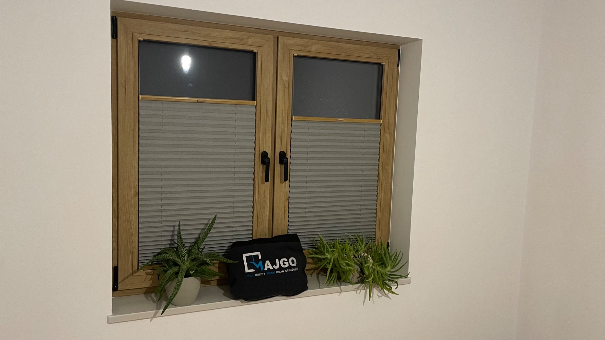 Zamknięte, szare plisy okienne w drewnianej ramie okna, z czarnymi uchwytami, na parapecie stoją dwie doniczki z zielonymi roślinami i czarna torba z logo firmy.