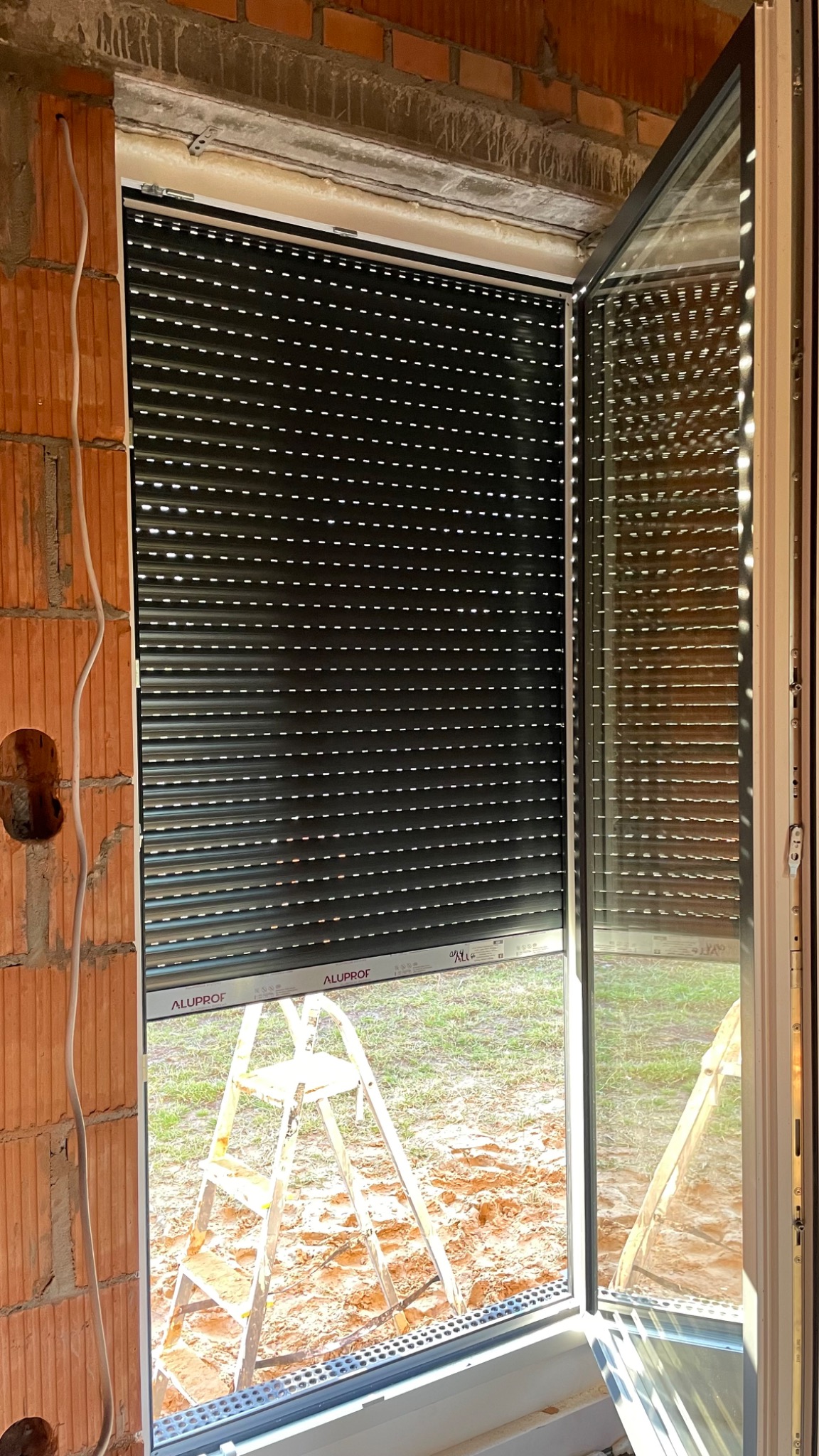 Montaż zewnętrznej rolety okiennej w surowym stanie, widoczna drabina, mur z cegły i wystające kable.