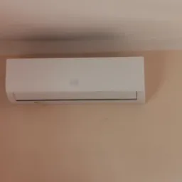 Biała jednostka klimatyzacji zamontowana na jasnoróżowej ścianie, widoczna z dołu pod lekkim kątem, z delikatnym cieniem na suficie.