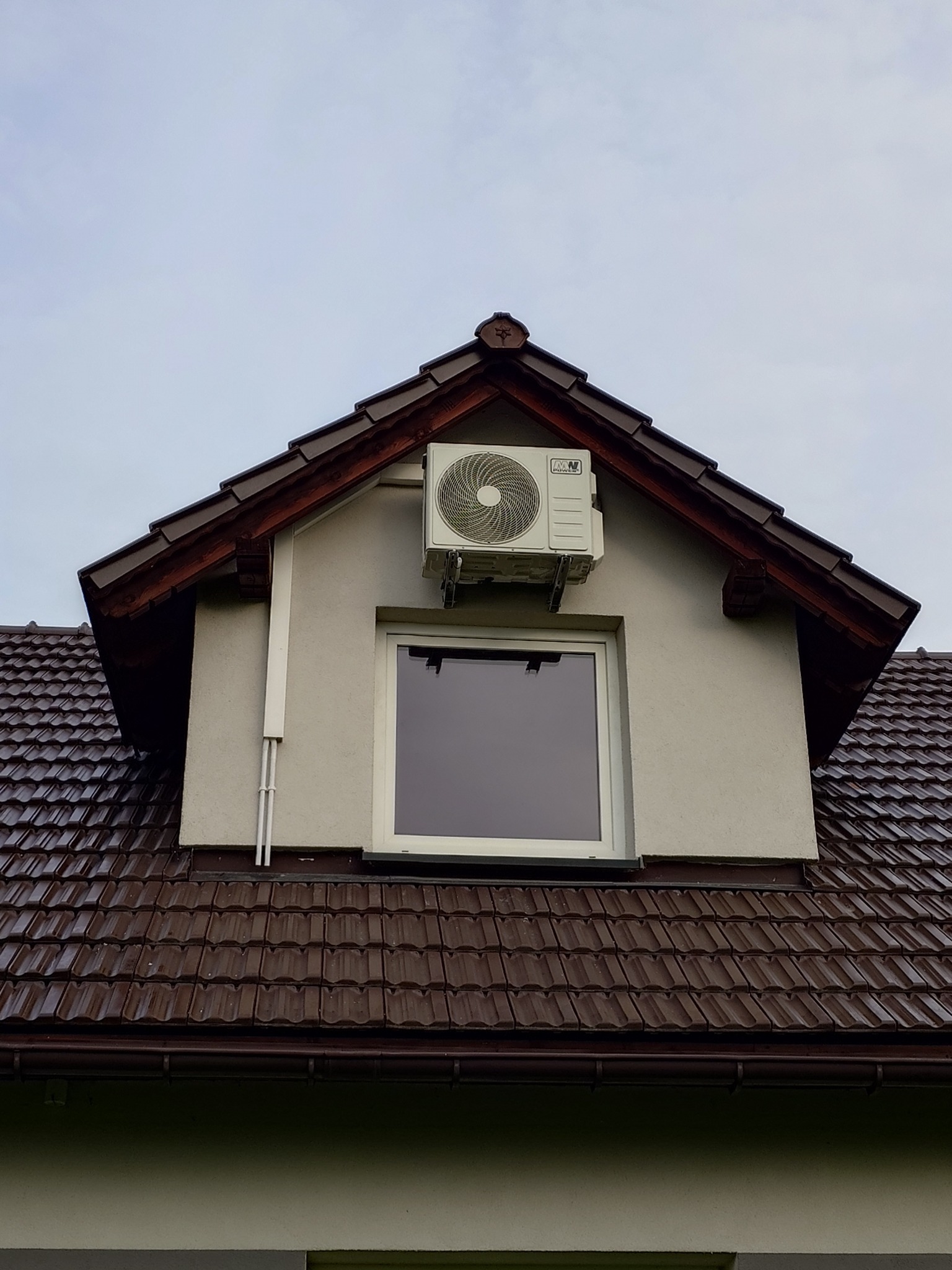 Zewnętrzna jednostka klimatyzatora zamontowana nad oknem na poddaszu domu z brązową dachówką, widoczne rury instalacyjne.