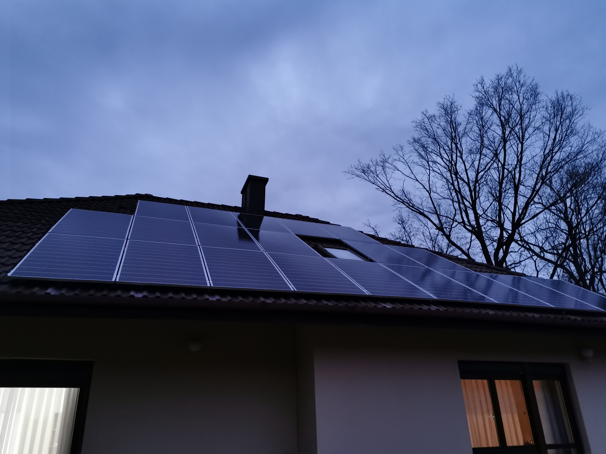 Panele słoneczne zamontowane na dachu domu jednorodzinnego, widoczne okno dachowe i komin, na tle pochmurnego nieba i drzew bez liści.