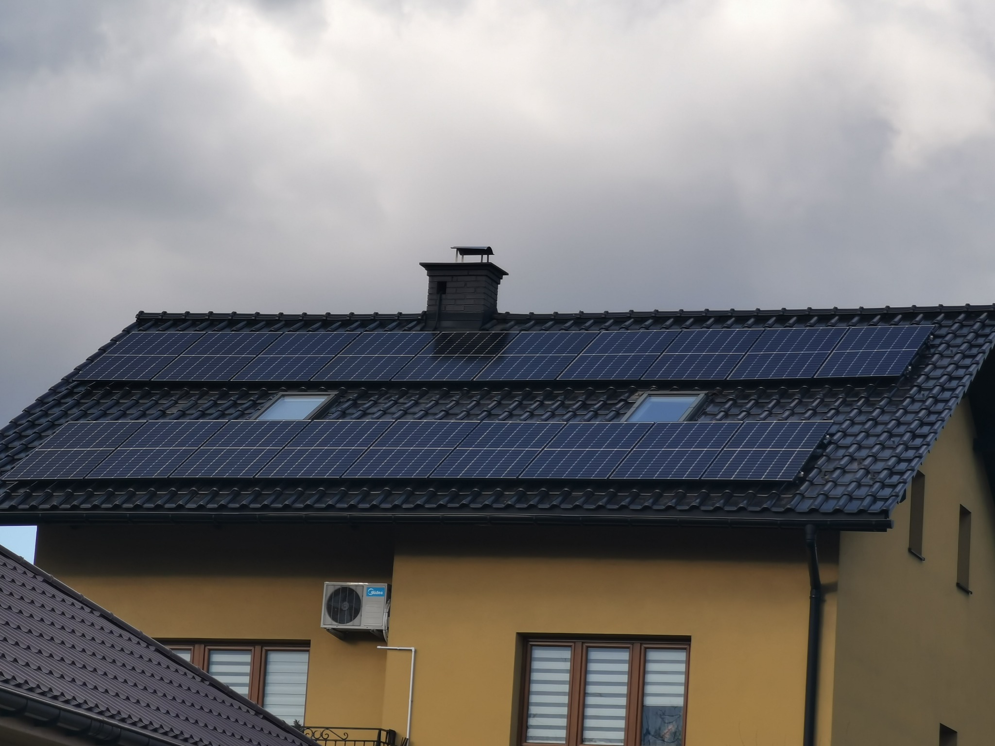 Dom z zamontowanymi panelami fotowoltaicznymi na dachu pokrytym czarną dachówką, widoczny komin i okna dachowe, na ścianie zewnętrzna jednostka klimatyzacji Midea, pochmurne niebo w tle.