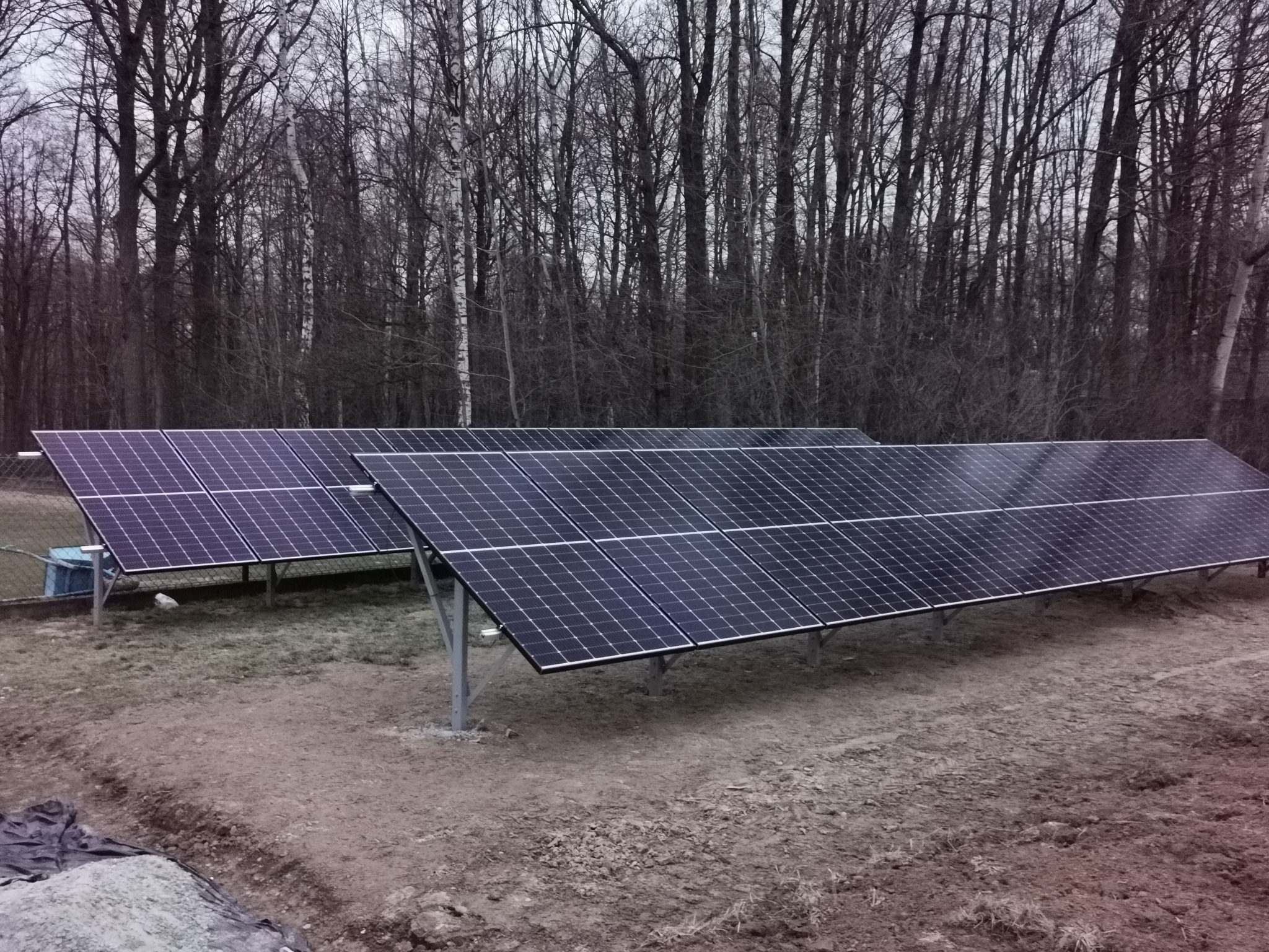 Dwa rzędy paneli słonecznych ustawione na metalowych stelażach na tle ponurego lasu bezlistnego, widoczne ślady prac ziemnych.
