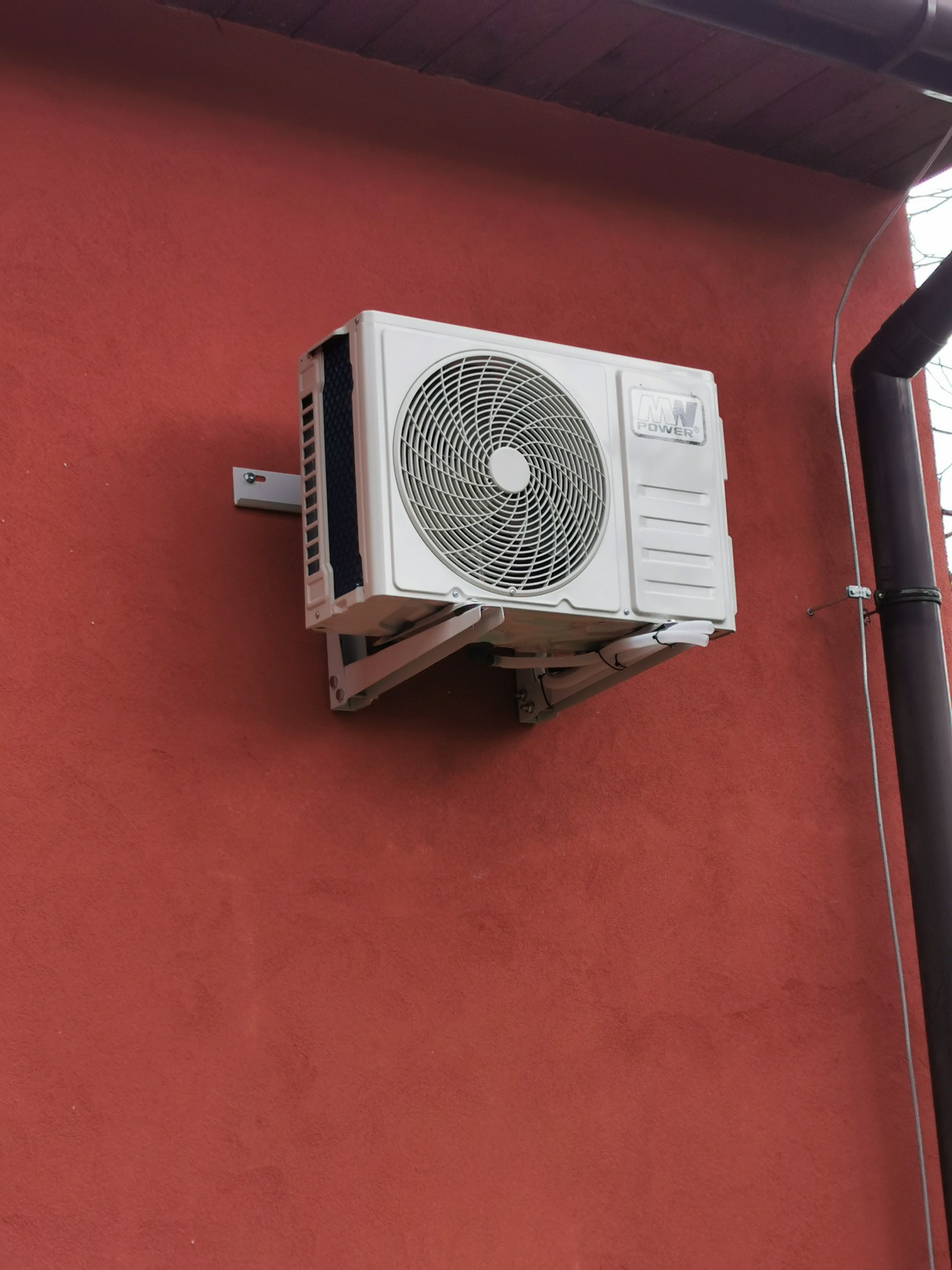 Biała jednostka zewnętrzna klimatyzatora zamontowana na czerwonej ścianie budynku, widoczne rury odprowadzające skropliny i brązowa rynna obok.