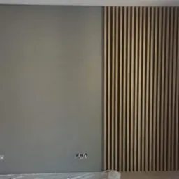 Ściana wewnętrzna z szarą, gładką powierzchnią i dekoracyjnym panelem z drewnianych listew, widoczne wystające kable elektryczne i okno z odbiciem samochodu.