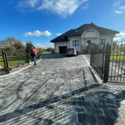 Ułożona kostka brukowa na podjeździe do domu jednorodzinnego z otwartą, kutą bramą wjazdową i furtką; widoczny fragment ogrodzenia, samochód i osoba idąca w kierunku domu.