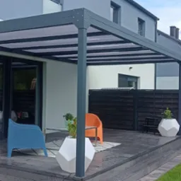 Zadaszony taras z szarą konstrukcją, drewnianą podłogą, nowoczesnymi donicami i kolorowymi krzesłami ogrodowymi, widok na ciemne drewniane ogrodzenie i budynki mieszkalne w tle.