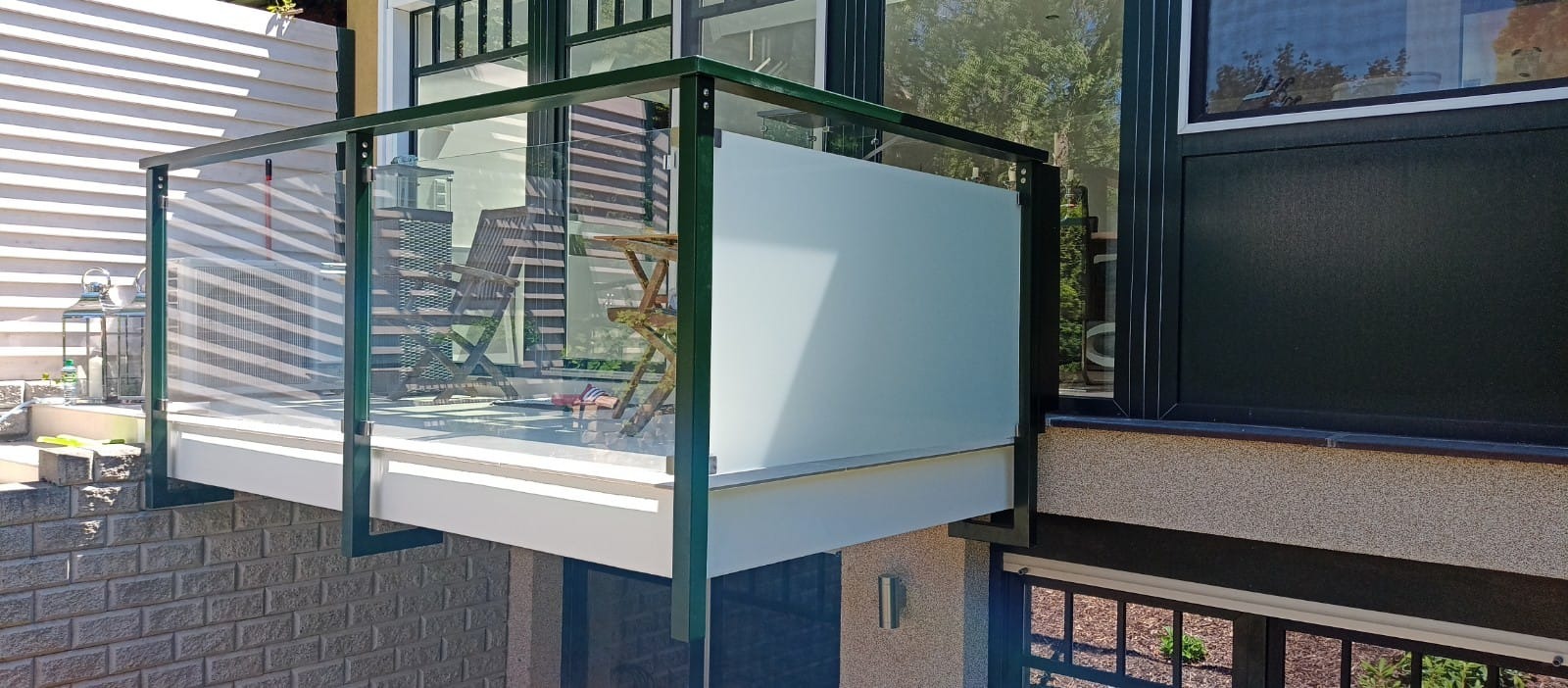 Nowoczesny balkon z balustradą ze szkła hartowanego i zielonymi metalowymi ramami, widok z dołu, częściowo zasłonięty matową szybą, meble ogrodowe w tle.