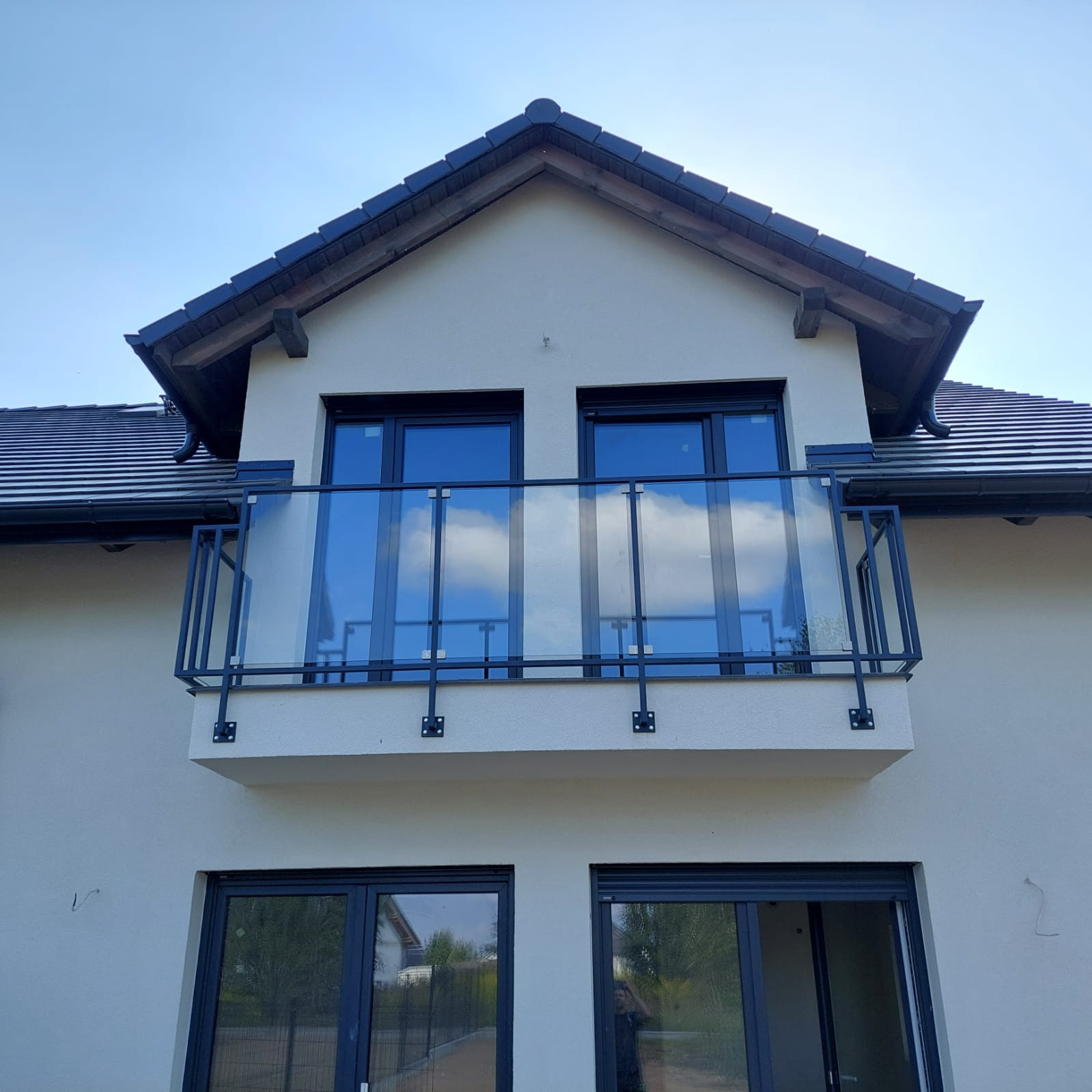 Balkon z nowoczesną balustradą szklaną z czarnymi, metalowymi elementami mocującymi, odbijający błękitne niebo z chmurami w szklanych taflach, na tle jasnej elewacji domu z ciemną dachówką.