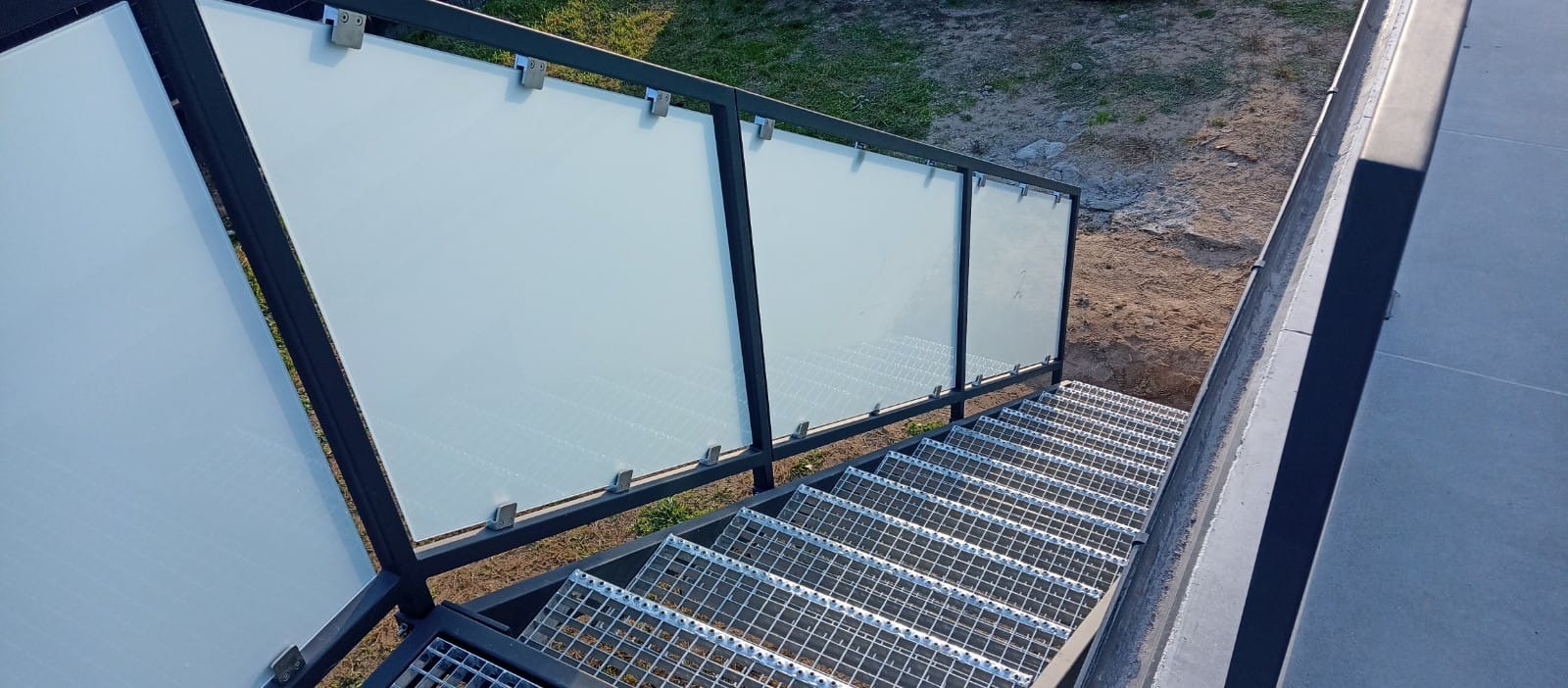 Metalowe schody z kraty pomostowej, zabezpieczone balustradą ze szklanymi panelami, mocowanymi metalowymi uchwytami, widok z góry na nierówny teren.