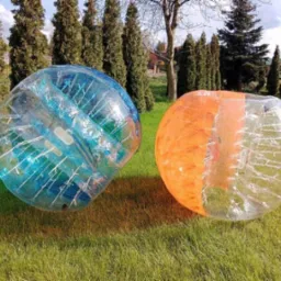 Dwie kule sferyczne: niebieska i pomarańczowa, wykonane z przezroczystego tworzywa, leżą na trawie w słoneczny dzień, w tle drzewa iglaste.