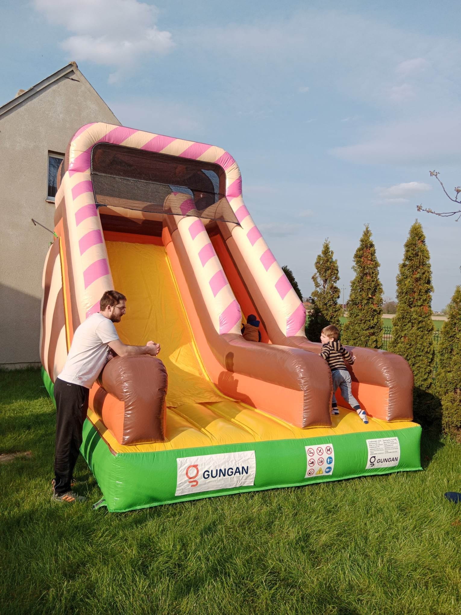 Nadmuchiwana zjeżdżalnia w pastelowych kolorach z motywem słodyczy, na której bawi się dziecko, a obok stoi dorosły mężczyzna, na tle zielonego trawnika i iglaków.