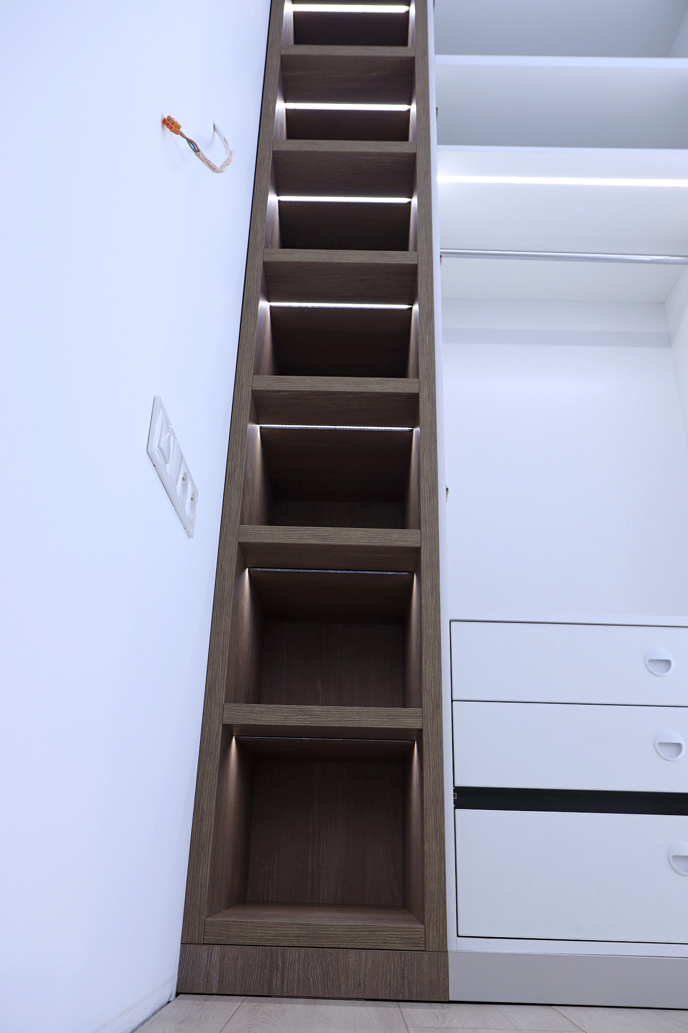 Nowoczesna szafa z wnękami oświetlonymi LED, przylegająca do białej ściany z gniazdkami elektrycznymi i wystającymi przewodami, obok biała szafa z szufladami.