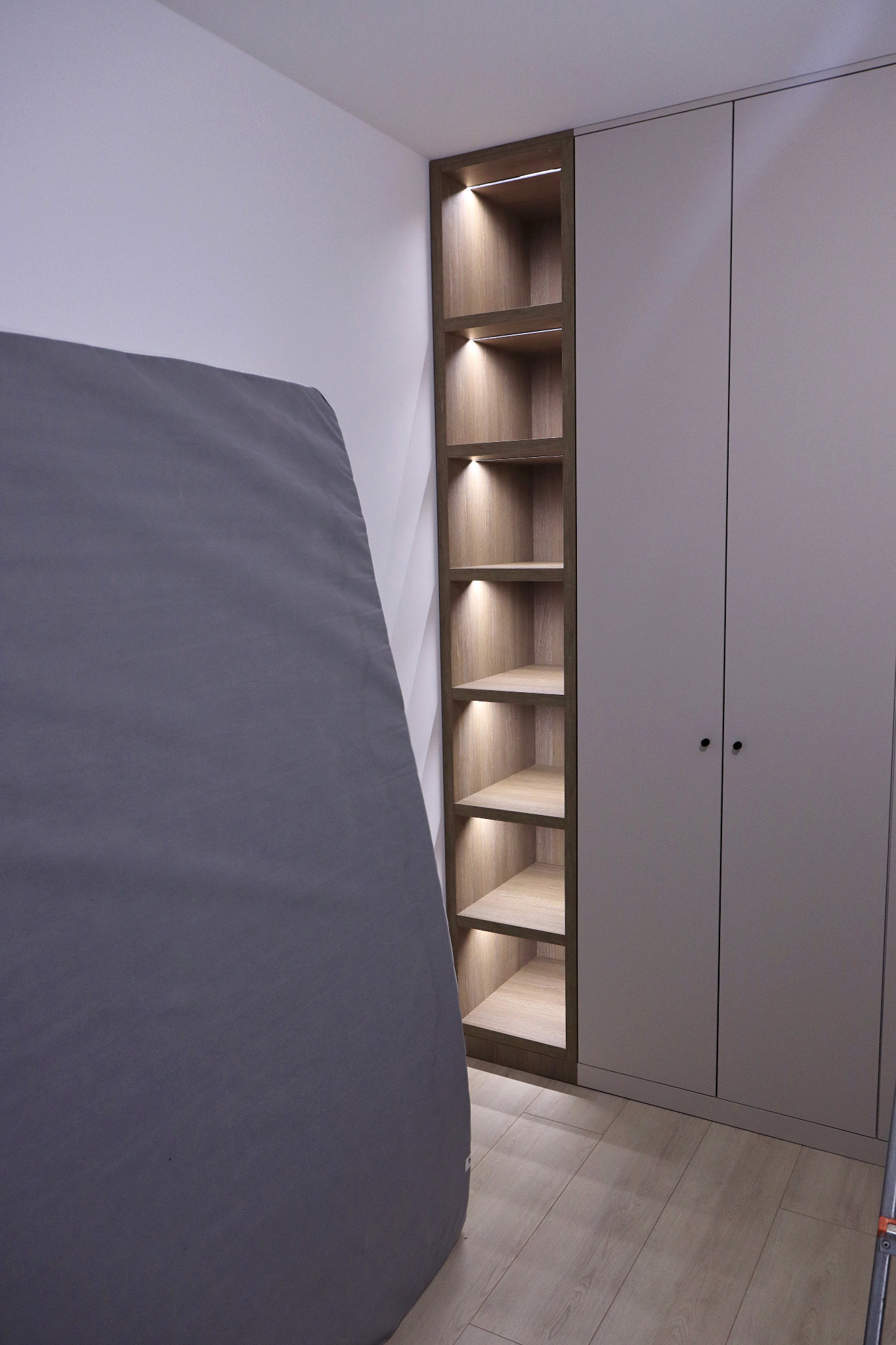 Szara szafa z wbudowanym regałem z podświetlanymi półkami, widoczny fragment materaca z boku.