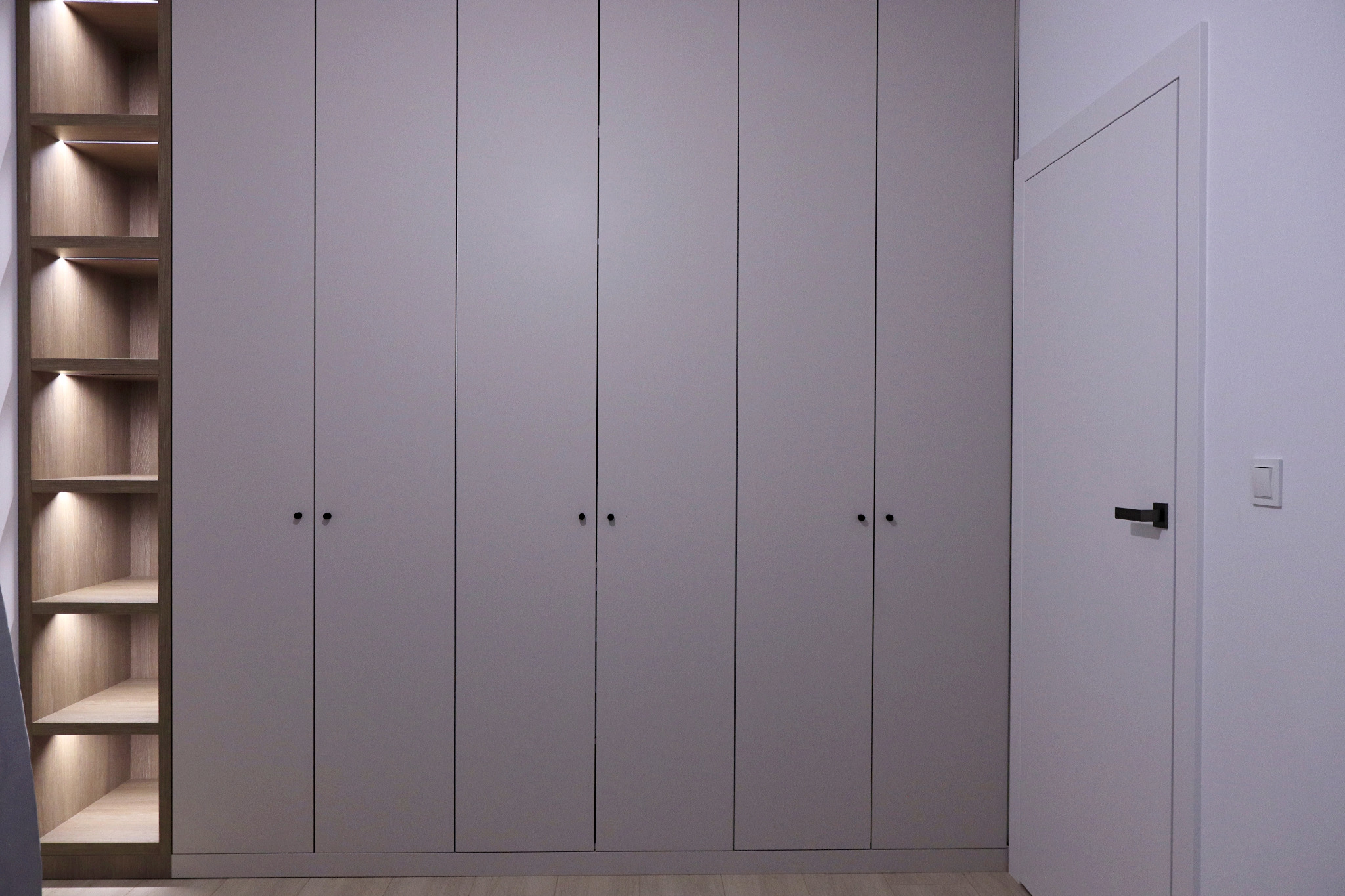 Szafa w zabudowie o gładkich, szarych frontach z minimalistycznymi, czarnymi uchwytami, z otwartym segmentem z podświetlonymi półkami po lewej stronie, przylegająca do białych drzwi z czarną klamką.