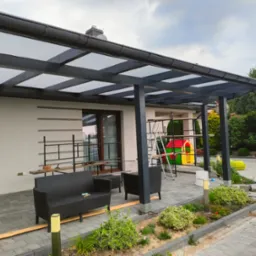 Zadaszony taras z drewnianą konstrukcją i przezroczystym pokryciem, w tle biały budynek i plac zabaw, widoczne meble ogrodowe i roślinność.