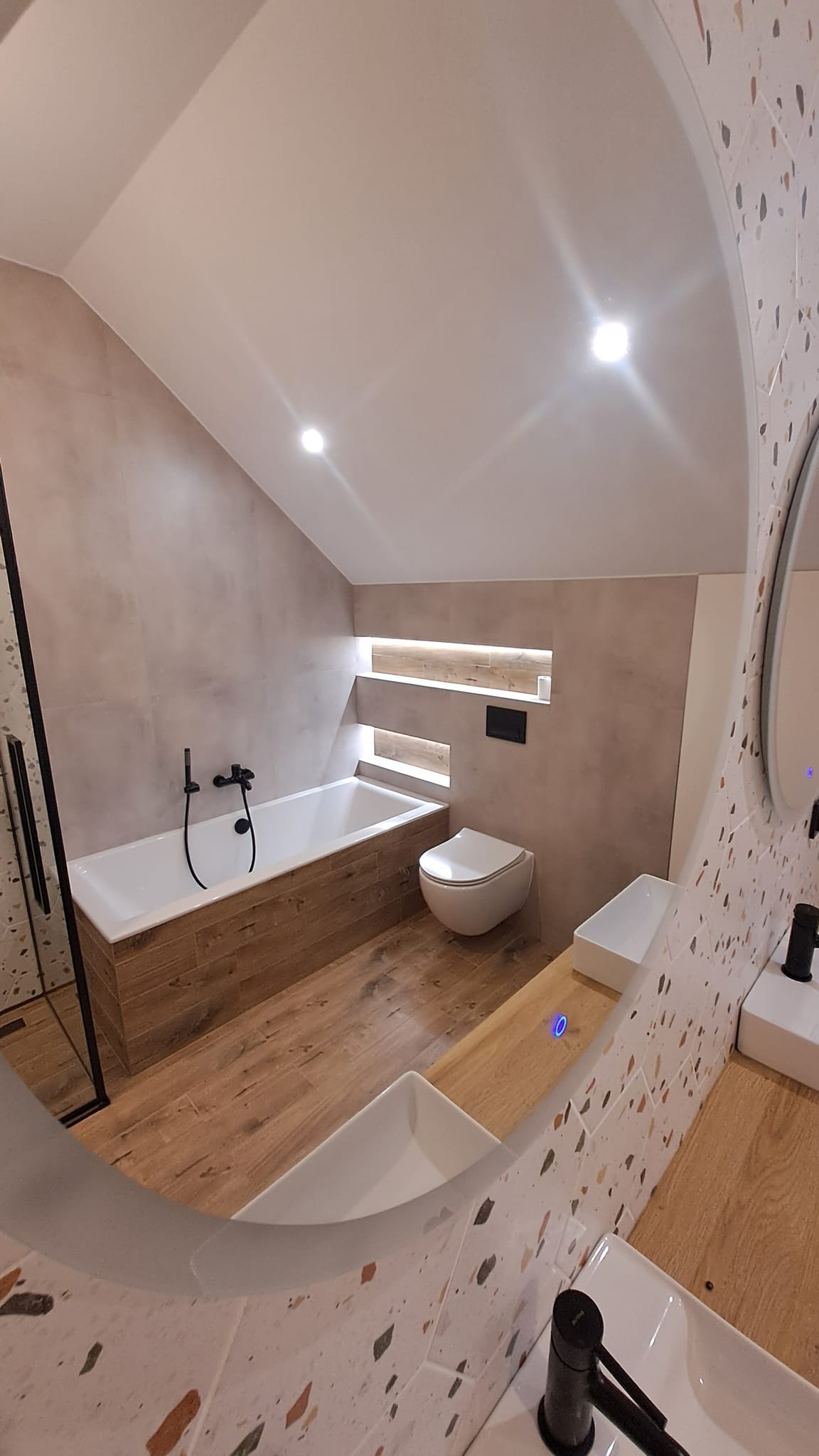 Nowoczesna łazienka na poddaszu z wanną obudowaną drewnem, toaletą podwieszaną i umywalkami na drewnianym blacie, widziana przez lustro z mozaikową ramą.