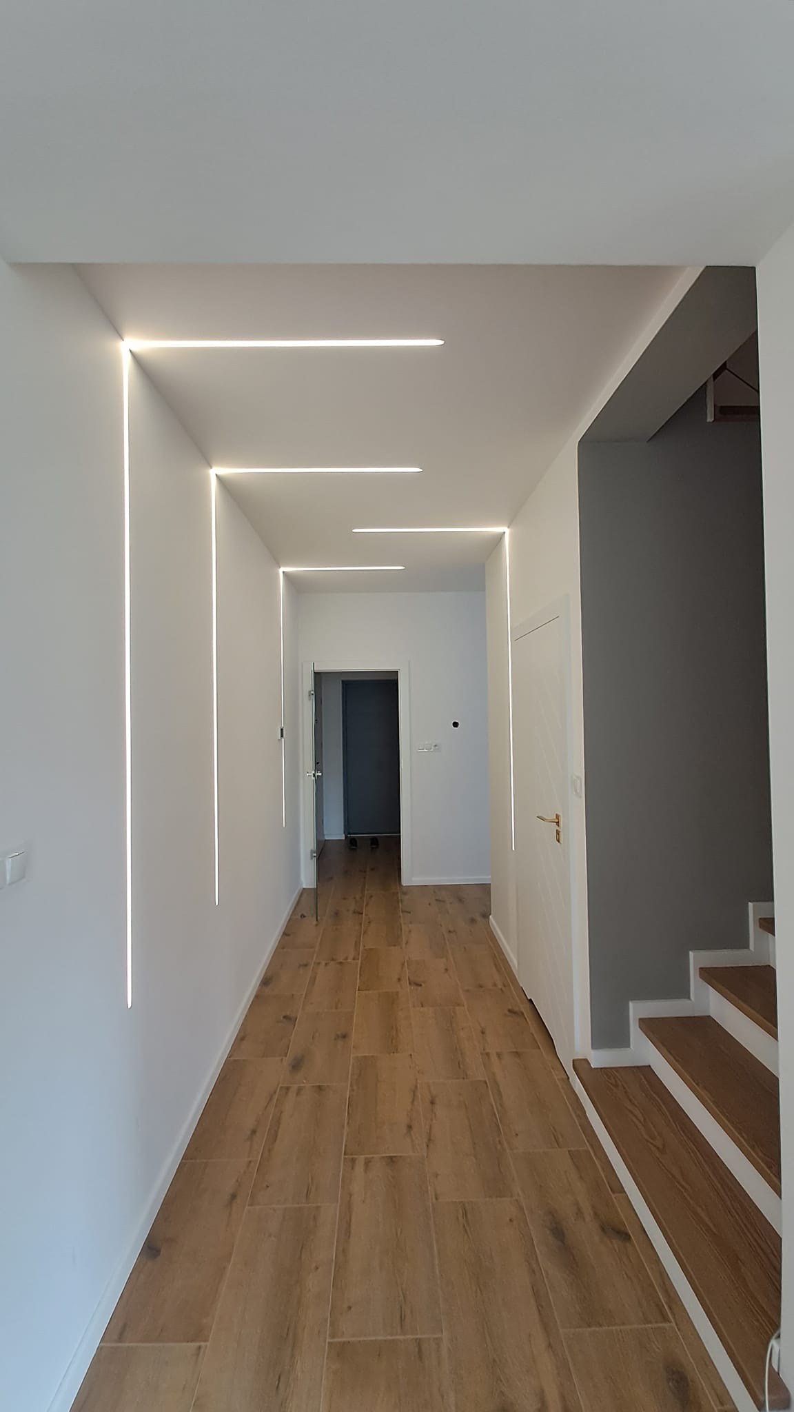 Nowoczesny korytarz z oświetleniem liniowym LED wpuszczonym w ściany i sufit, drewnopodobna podłoga z płytek, widoczne schody z szarą ścianą.