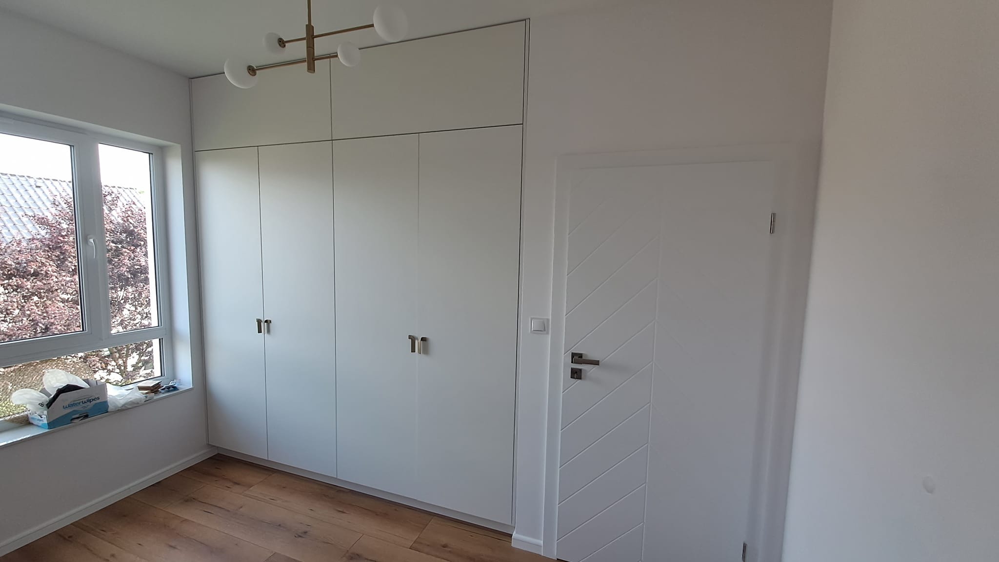 Biała zabudowa szafy z górnymi szafkami, minimalistyczny żyrandol z kulistymi kloszami, drzwi z frezowanym wzorem w jodełkę, widok z okna na drzewo, drewniana podłoga.