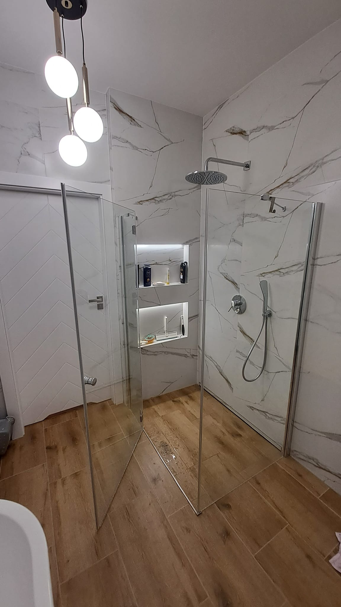 Nowoczesna kabina prysznicowa walk-in z marmurowymi płytkami i drewnopodobną podłogą we Wrocławiu, z wnękami podświetlonymi LED na kosmetyki.