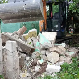 Pomarańczowy minikoparka CASE z szarą łyżką nad stertą gruzu betonowego z resztkami farby, na tle zielonej siatki i drzew