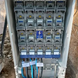 Otwarta skrzynka rozdzielcza z zamontowanymi bezpiecznikami i podłączonymi kablami, etykiety z opisem kabli przymocowane do przewodów, widok z góry na skrzynkę umieszczoną w ziemi.