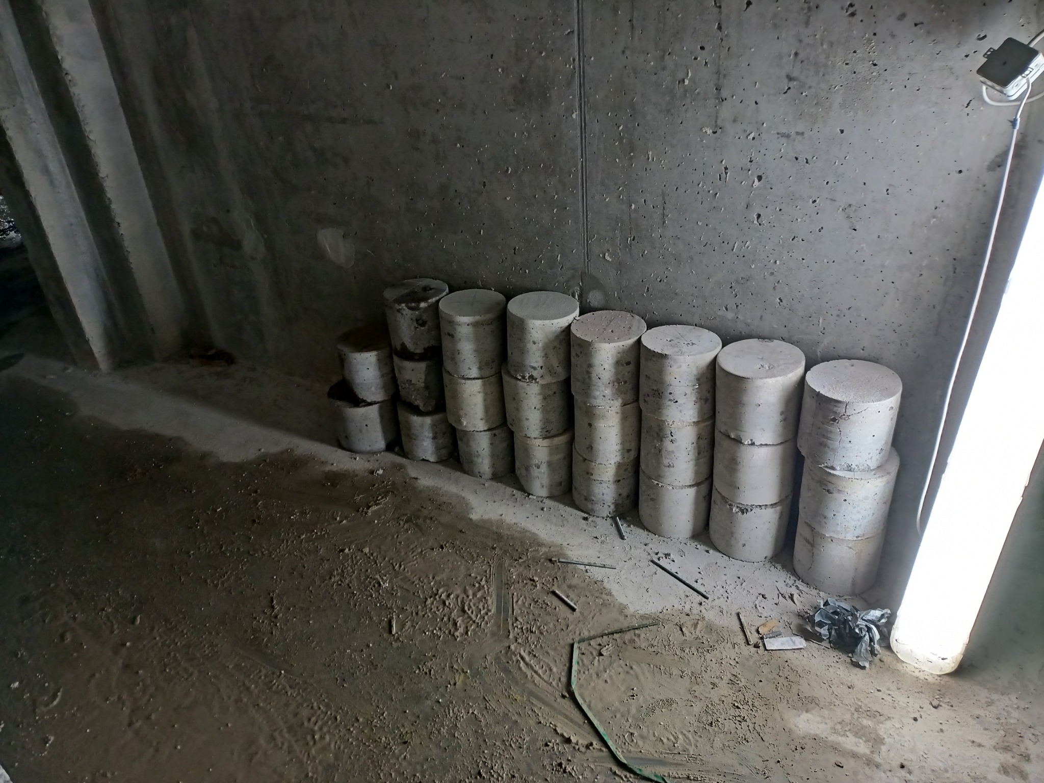Cylindryczne próbki betonu ułożone w stosy na podłodze w surowym wnętrzu, widoczne ślady wody i zabrudzenia na podłożu, w tle betonowa ściana z widoczną instalacją elektryczną.