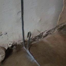 Damian Figielek - Metalowa listwa przypodłogowa z haczykowatym zakończeniem przymocowana do nierównej, popękanej ściany w trakcie montażu.