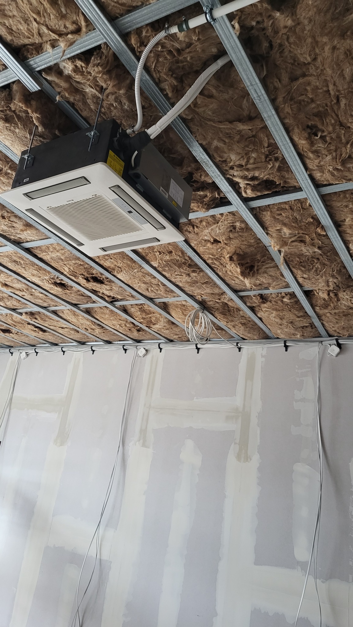 Kasetonowy klimatyzator zamontowany w suficie podwieszanym z izolacją z wełny mineralnej, widoczne przewody i elementy konstrukcji sufitu.
