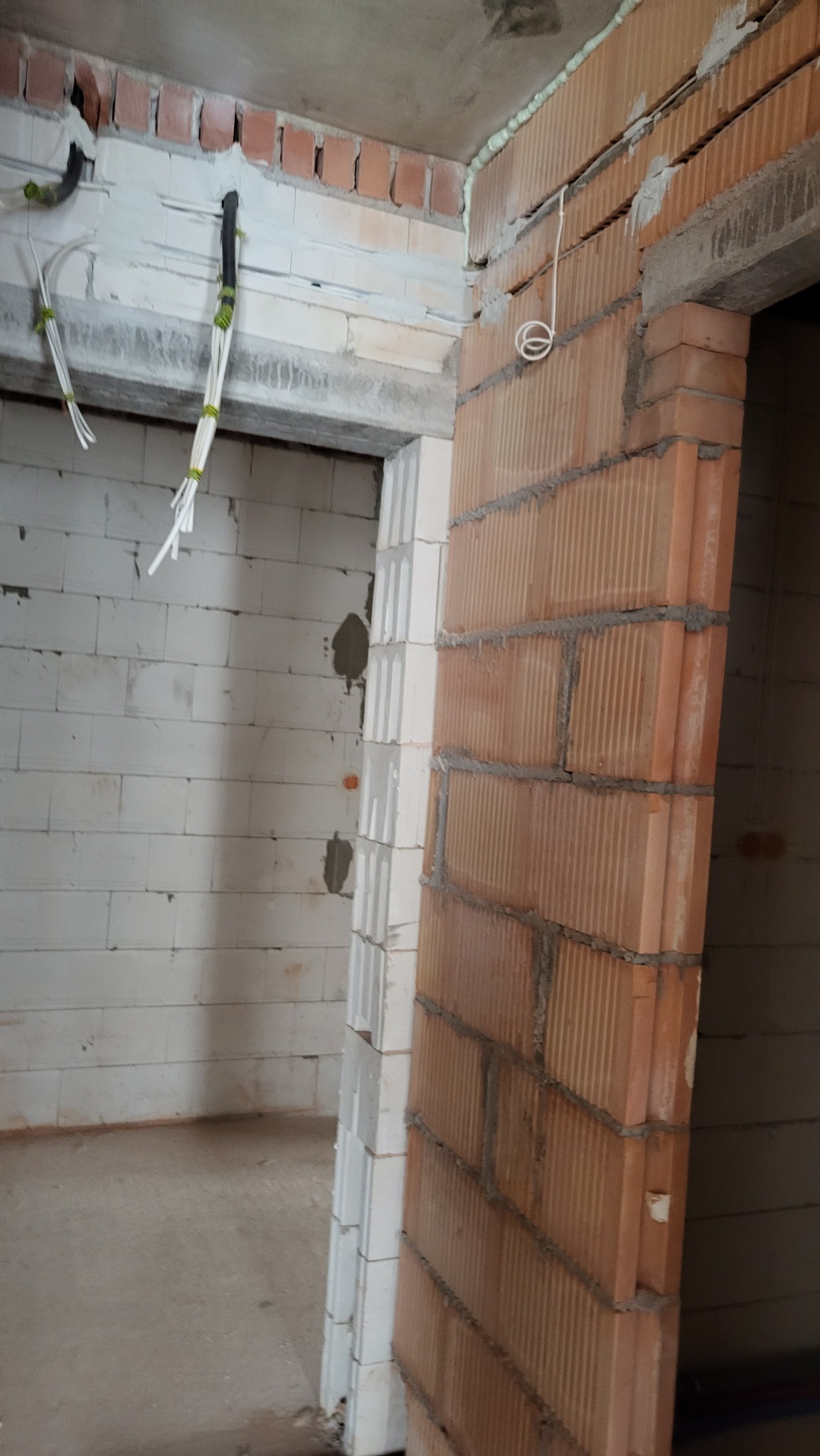 Surowy stan wnętrza z instalacją elektryczną: przewody w peszlach przymocowane do ściany z białego bloczka, widoczna wiązka kabli i fragment murowanej futryny z czerwonej cegły dziurawki.