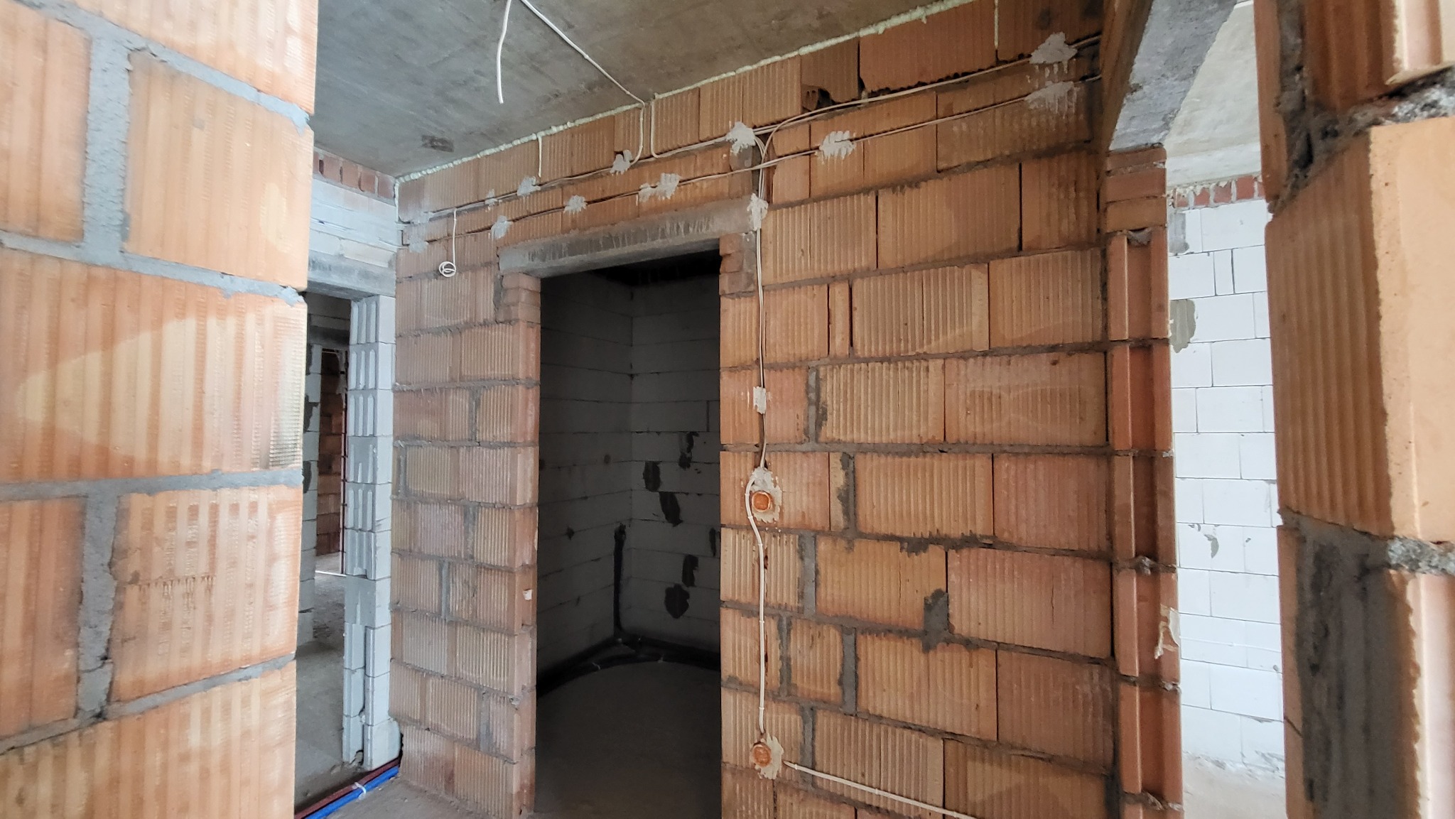 Surowy stan wnętrza w trakcie prac wykończeniowych: ściany z cegły ceramicznej z widocznymi przewodami elektrycznymi zamocowanymi do ściany, otwór drzwiowy bez ościeżnicy, fragment widocznego...