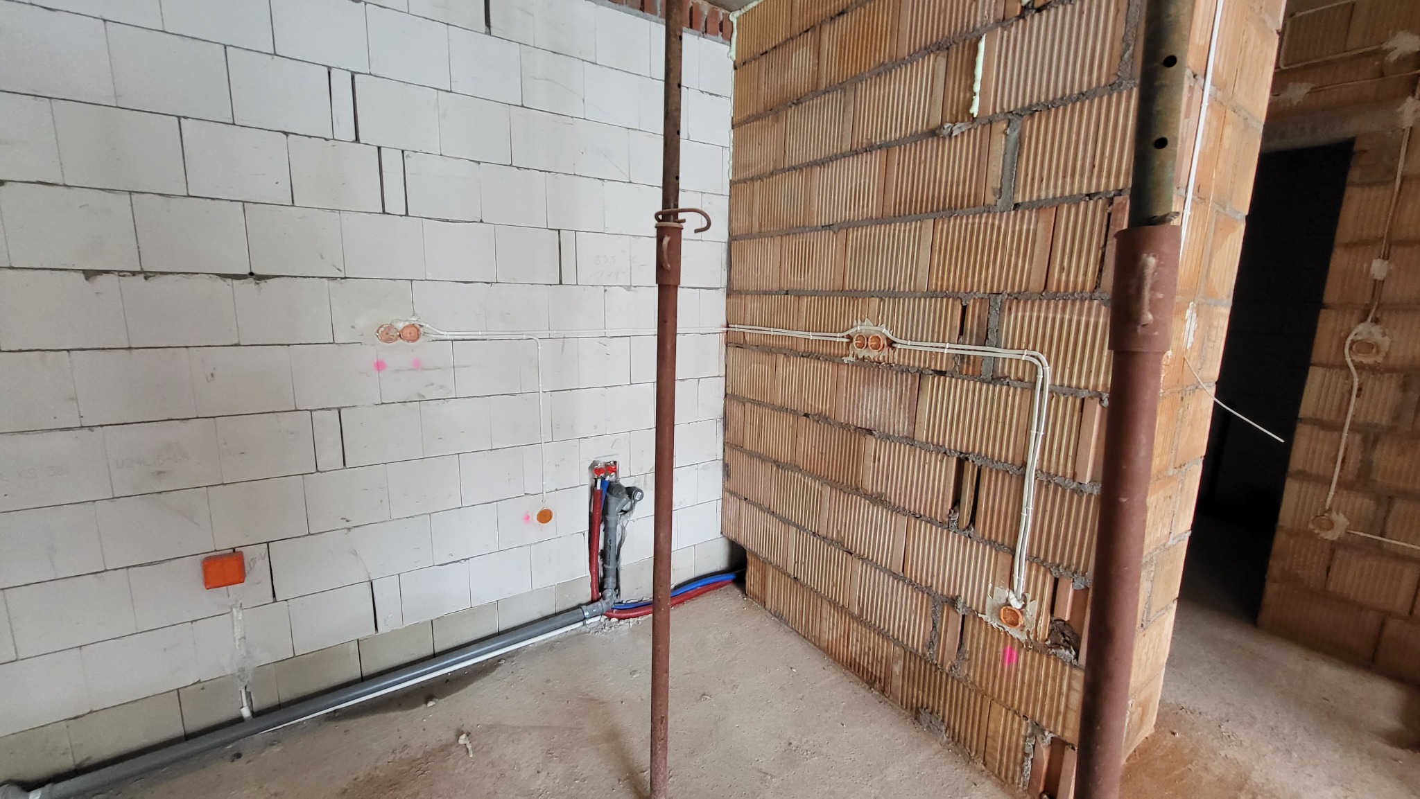 Wnętrze w stanie surowym: białe ściany z betonu komórkowego i ściany z czerwonej cegły, widoczne puszki elektryczne, rury instalacyjne i metalowa podpora stropowa.