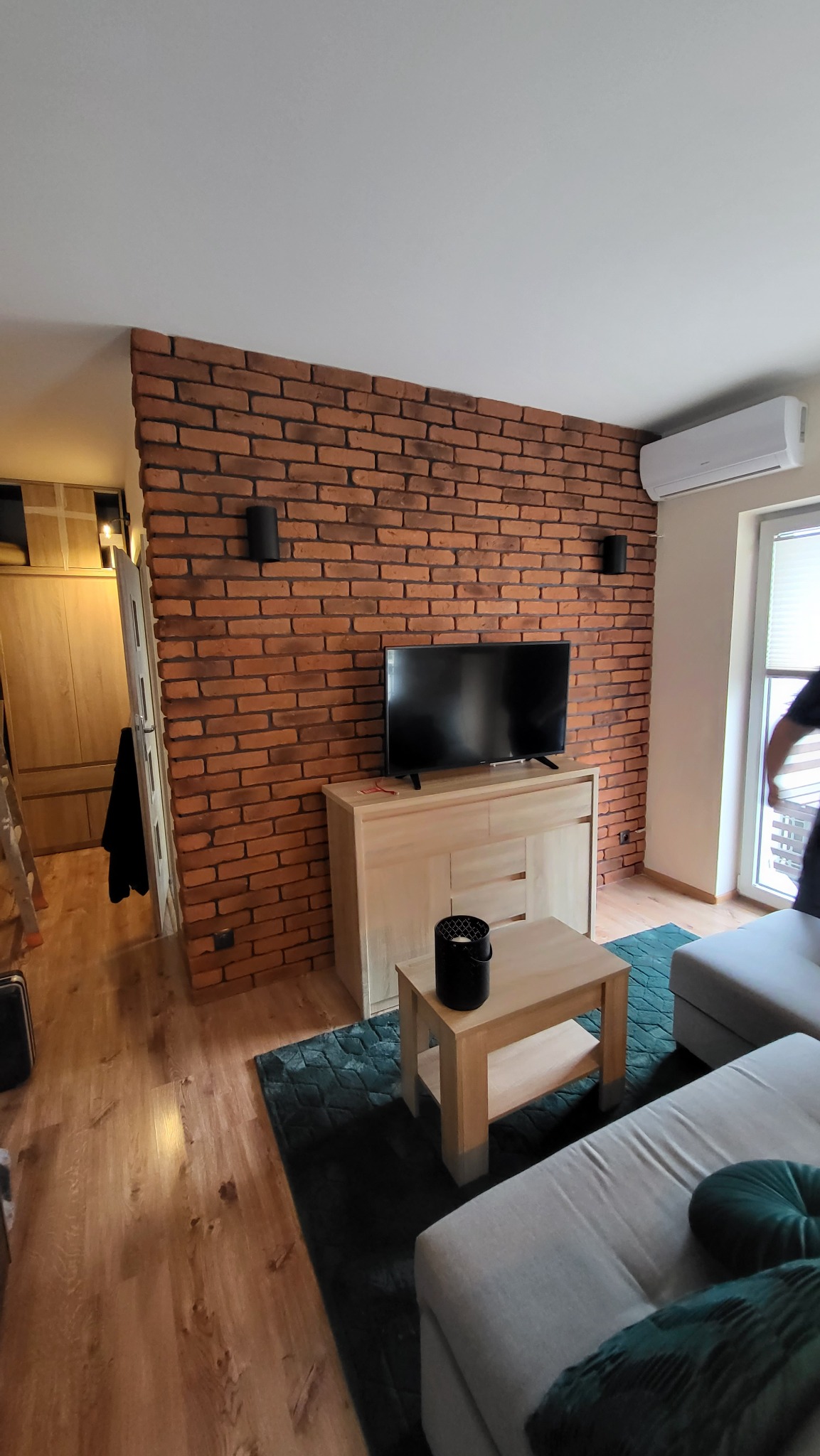 Salon z ceglaną ścianą, oświetleniem punktowym, telewizorem na komodzie i jasną sofą na zielonym dywanie.