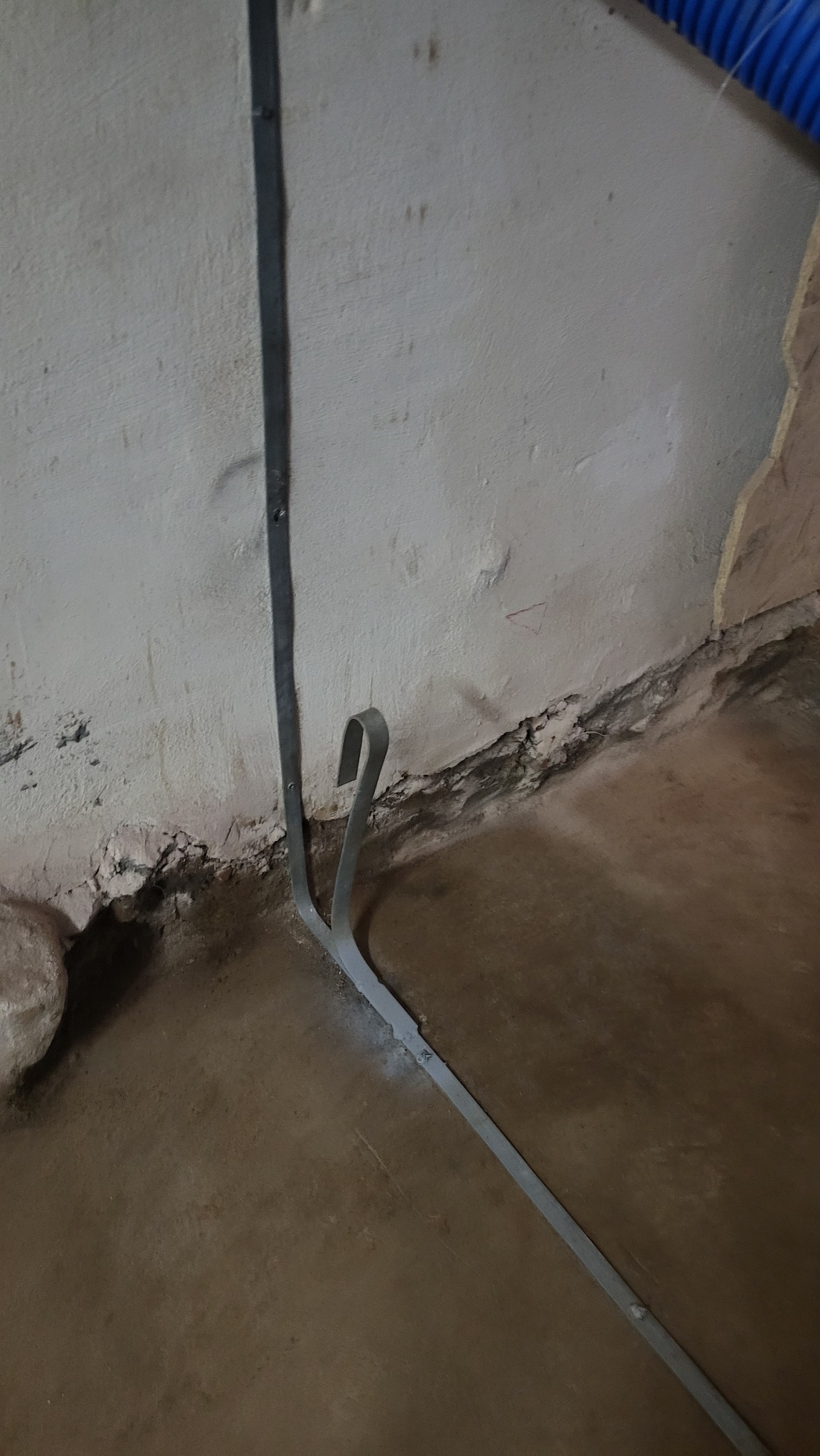 Metalowa listwa przypodłogowa z haczykowatym zakończeniem przymocowana do nierównej, popękanej ściany w trakcie montażu.