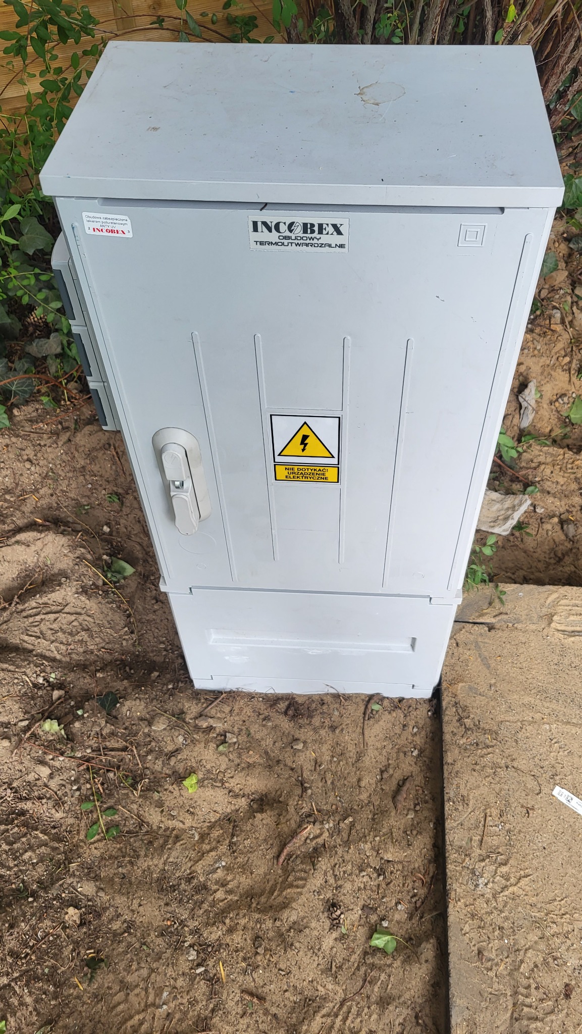 Szara, termoutwardzalna obudowa elektryczna IncoBex z ostrzeżeniem o napięciu, umieszczona na ziemi przy betonowej krawędzi, widok z góry.