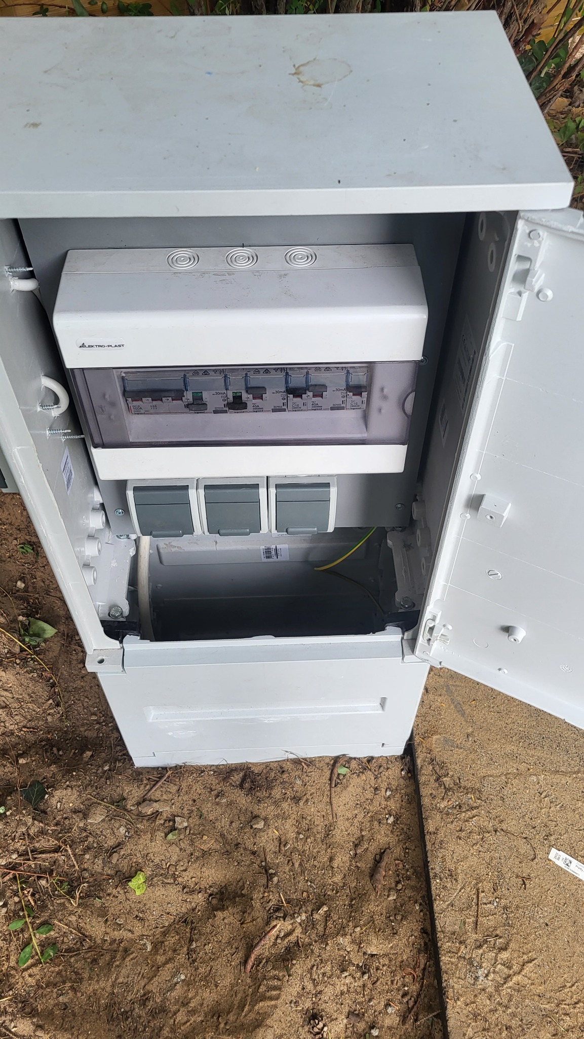 Otwarta, biała skrzynka elektryczna z widocznymi bezpiecznikami i przewodami, umieszczona na ziemi.